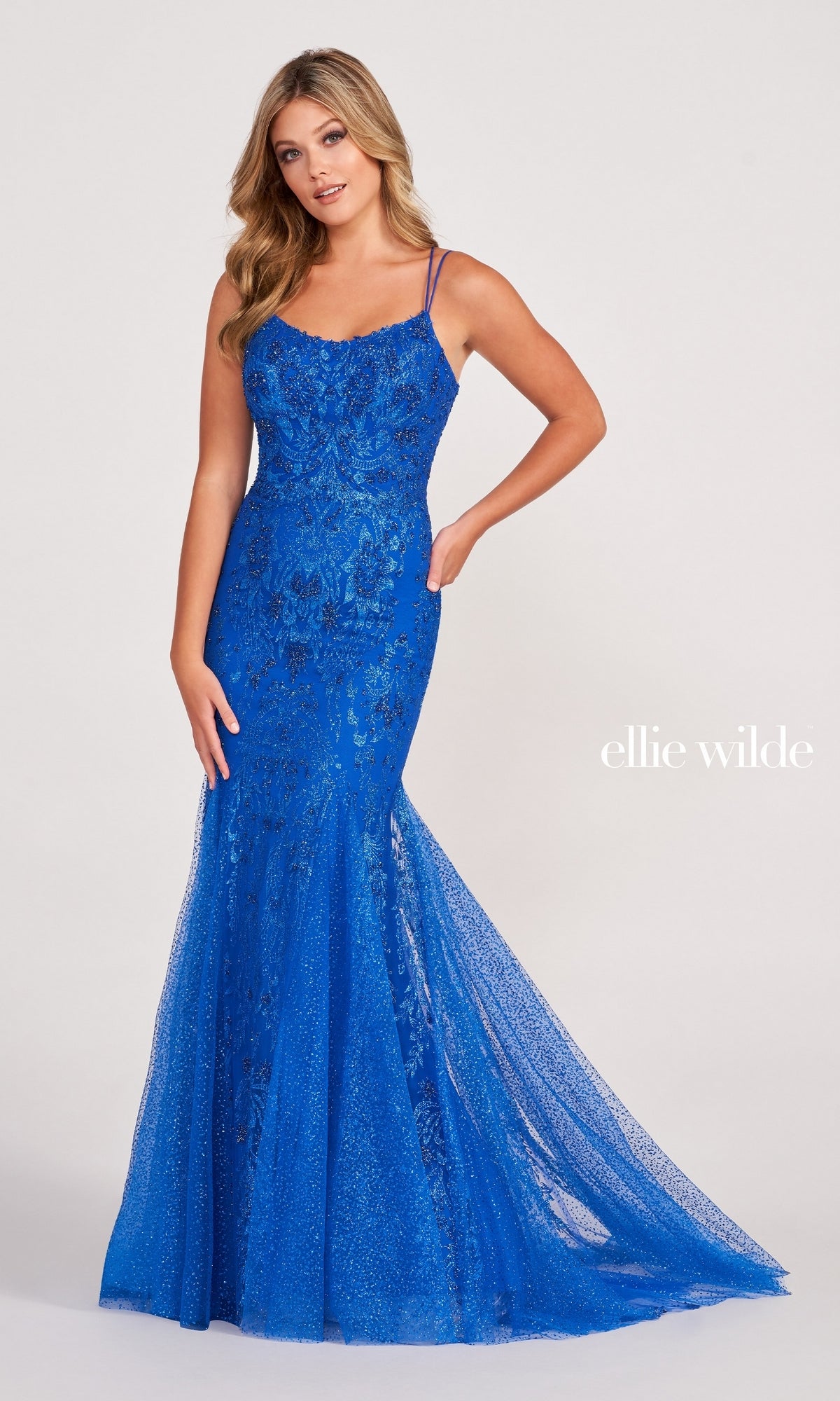 A woman poses against a plain background in the Glitter-Jersey Long Mermaid Ellie Wilde Prom Gown, a fitted royal blue dress with lace embellishments, spaghetti straps, a flared hem, and a subtle shimmer.